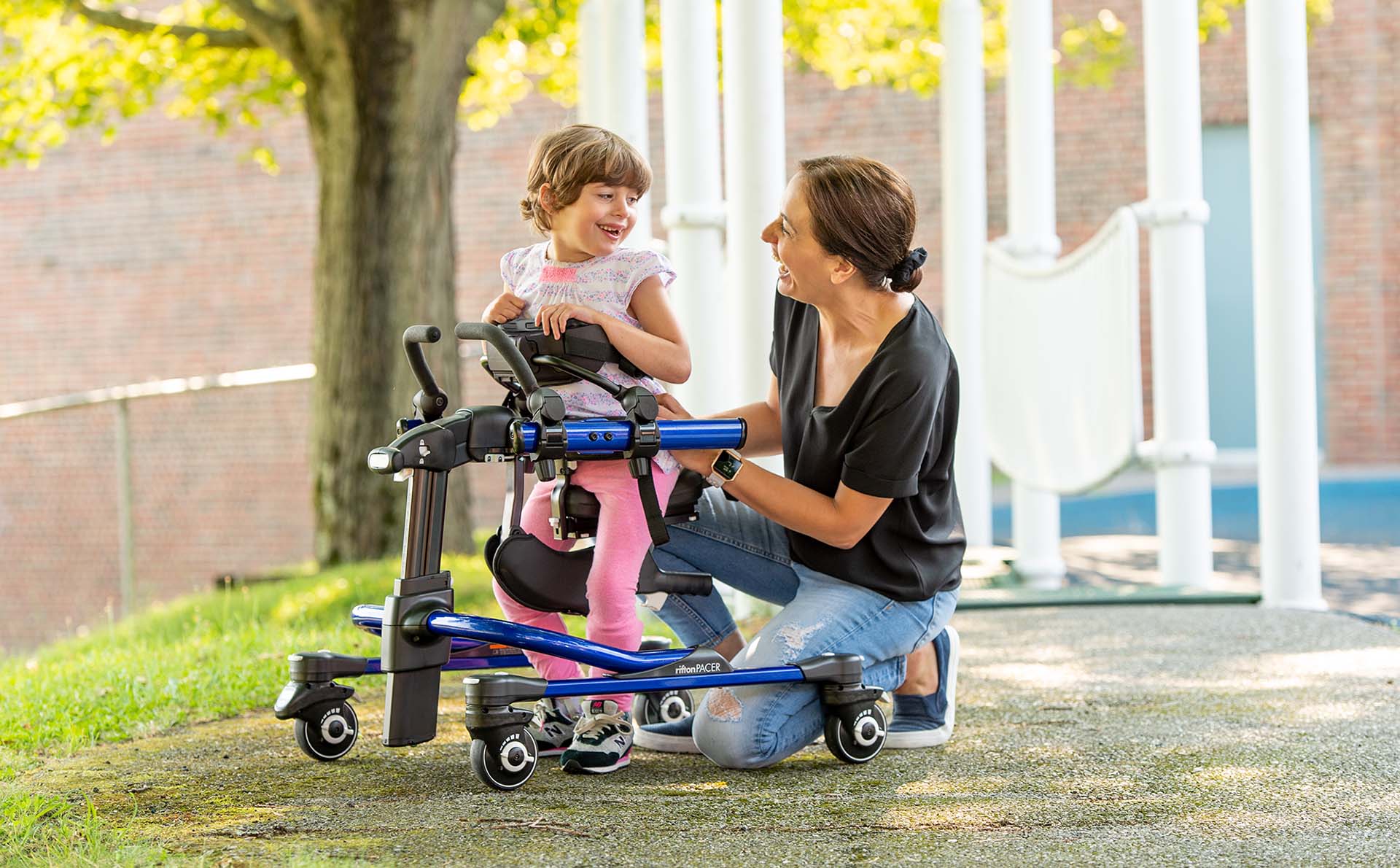 A little girl stands in a Rifton Stander and smiles at her caretaker.