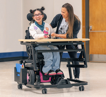 A girl sits at a Rifton Adaptive Desk and smiles at her caretaker.
