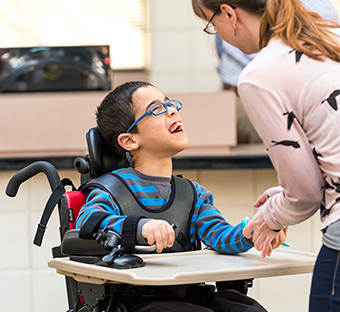 A boy sits in a Rifton Activity Chair and holds onto a Rifton Anchor and talks to his caretaker.