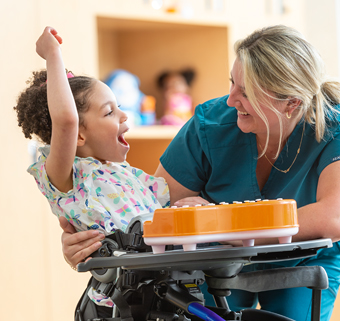 A girl stands in a Size 1 Rifton Stander and waves.