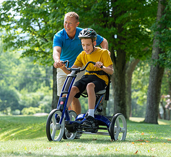 A boy rides on a Rifton Adaptive Tricycle with assistance from his therapist.