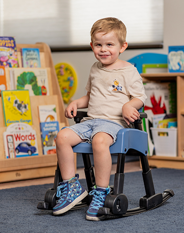 A boy sits on a Rifton Compass Chair with a rocking chair base and smiles.