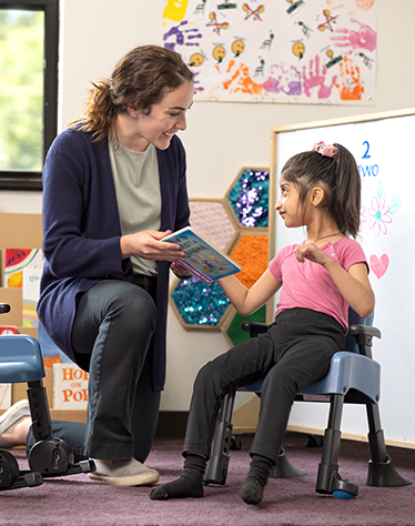 A girl sits on a Rifton Compass Chair and reads a book with her therapist.