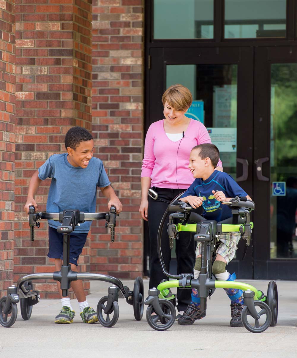 Two boys use Rifton Pacer gait trainers in front of a school while a physical therapist looks on.