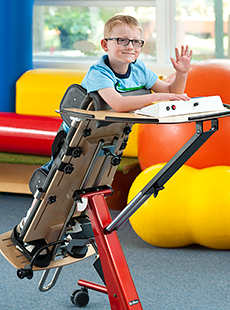 A small boy waves for the camera while standing in a Rifton Prone stander