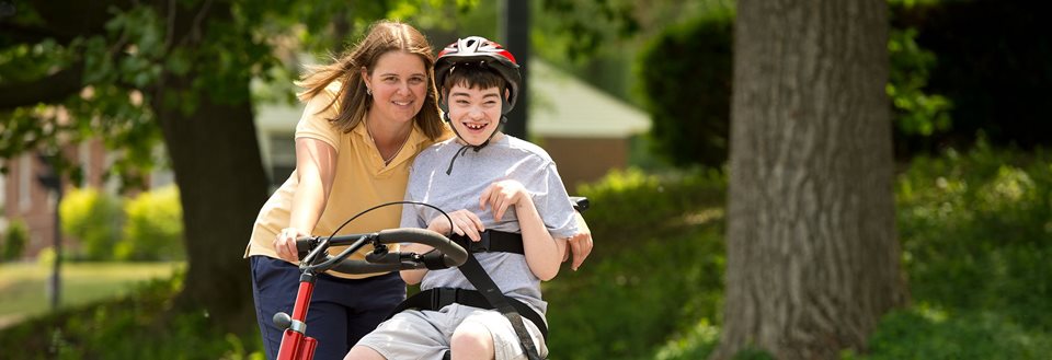 A school-aged boy rides a Rifton Adaptive Tricycle while his caregiver helps steer from behind using the rear steering bar