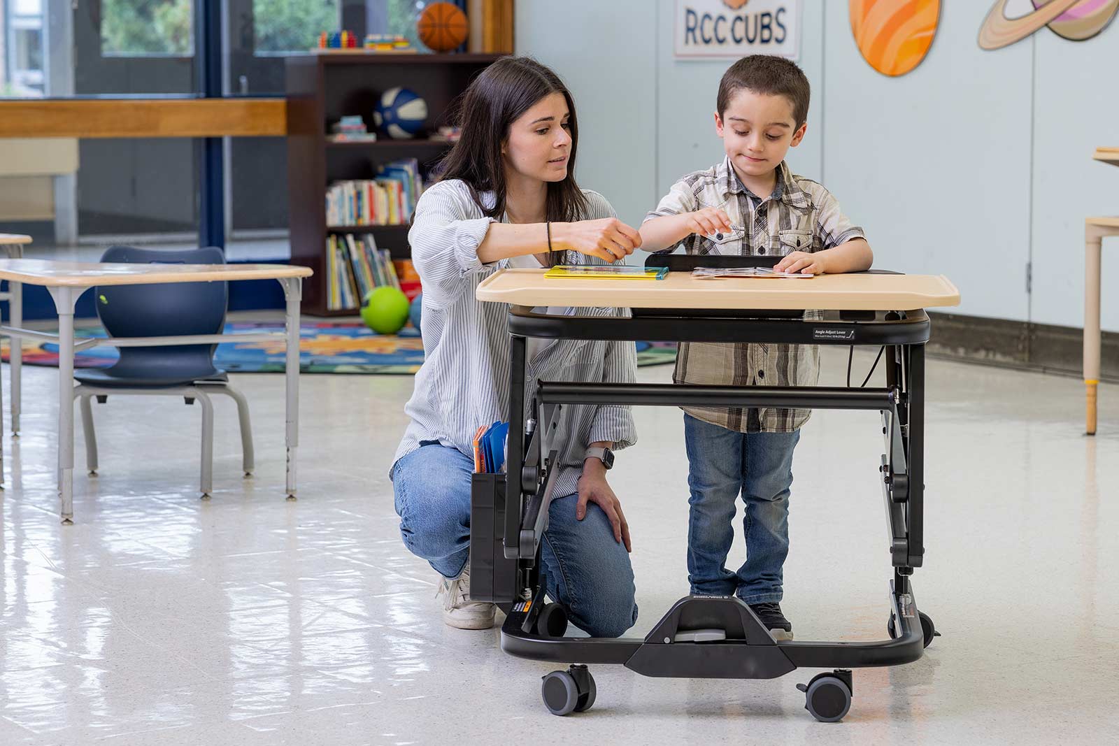 A boy stands by a Rifton Adaptive Desk and works in an activity book with his caretaker.