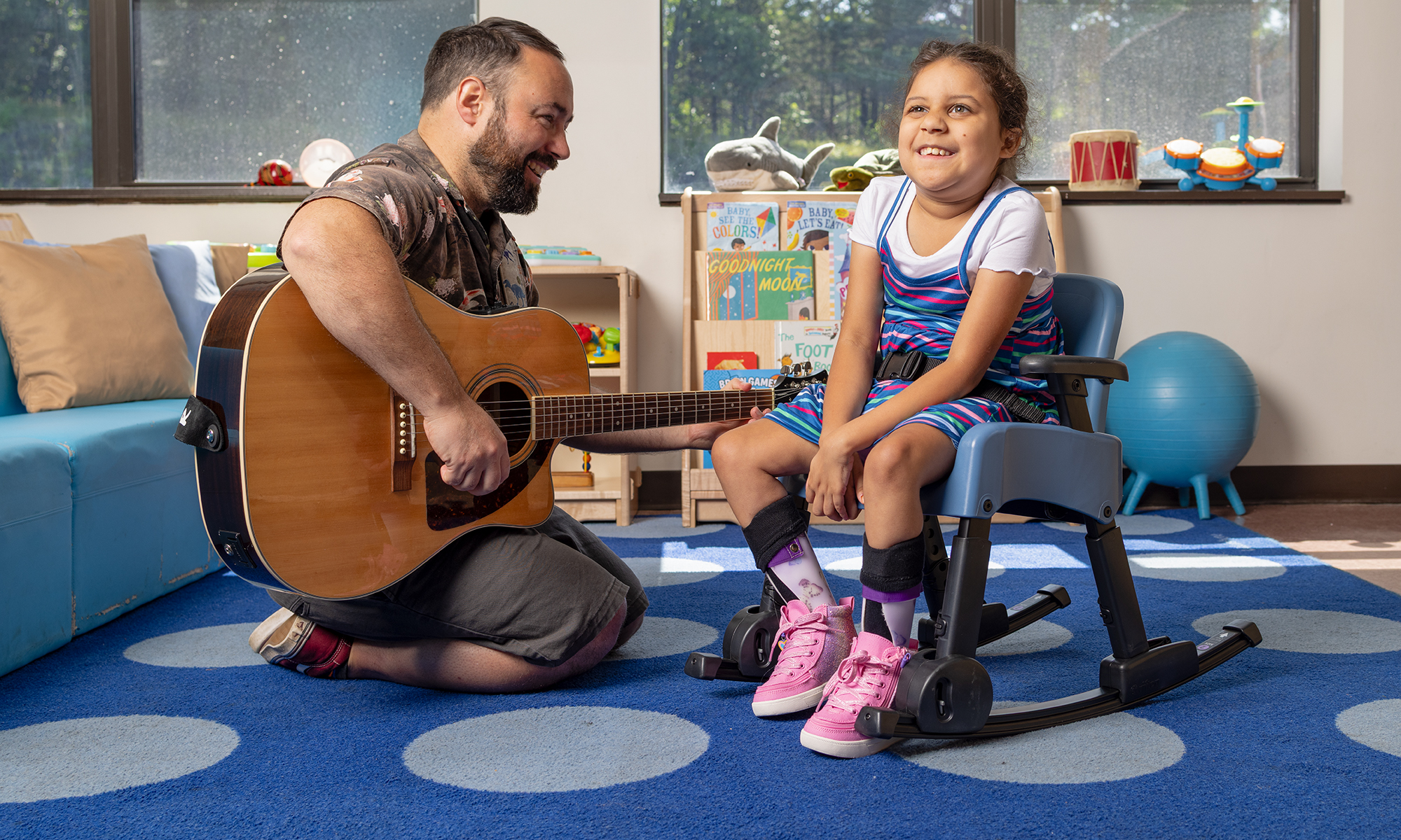 A girl sits in a Rifton Compass Chair with a rocking chair base and smiles at her music teacher, who is playing guitar.