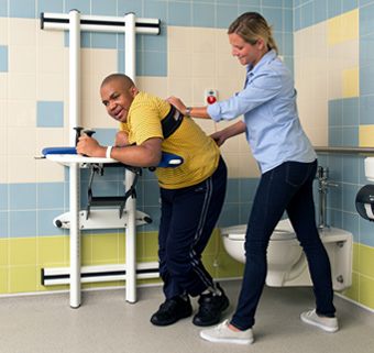 A young man in an orange striped shirt stands at a Rifton Support Station located in a bathroom.