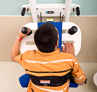 A young man in an orange striped shirt stands at a Rifton Support Station located in a bathroom.