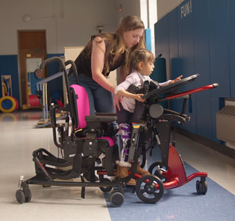 A physical therapists assists a young girl to stand in a Rifton prone stander from a Rifton Activity Chair.