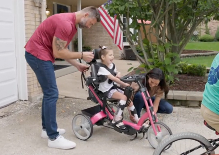 A mother and father help their daughter get on a Rifton Tricycle.