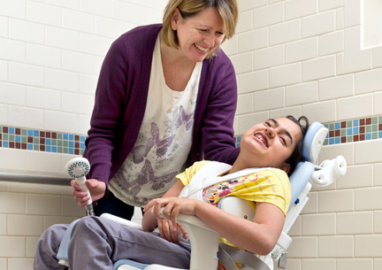 A girl sits on a Rifton HTS chair in a shower and smiles at her caretaker.