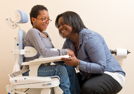 A young woman sits on a Rifton HTS and smiles at her caretaker.
