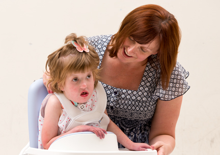 A girl sits on a Rifton HTS while here caregiver smiles at her.