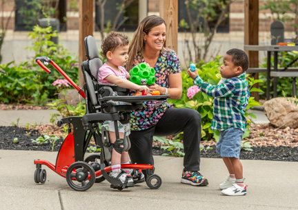 A little girl stands in a Rifton Stander and plays with a little boy while their caretaker watches.