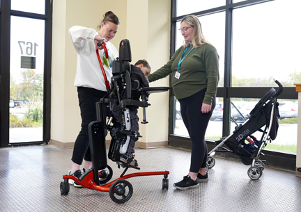 A boy stands in the Rifton Stander with the help of two therapists.