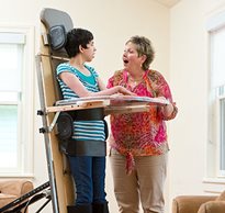 A caregiver laughs with a young teenage client who is standing in a Rifton Supine Stander.