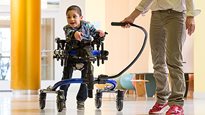A little boy walking in a blue Rifton dynamic Pacer gait trainer, with a caregiver helping him steer